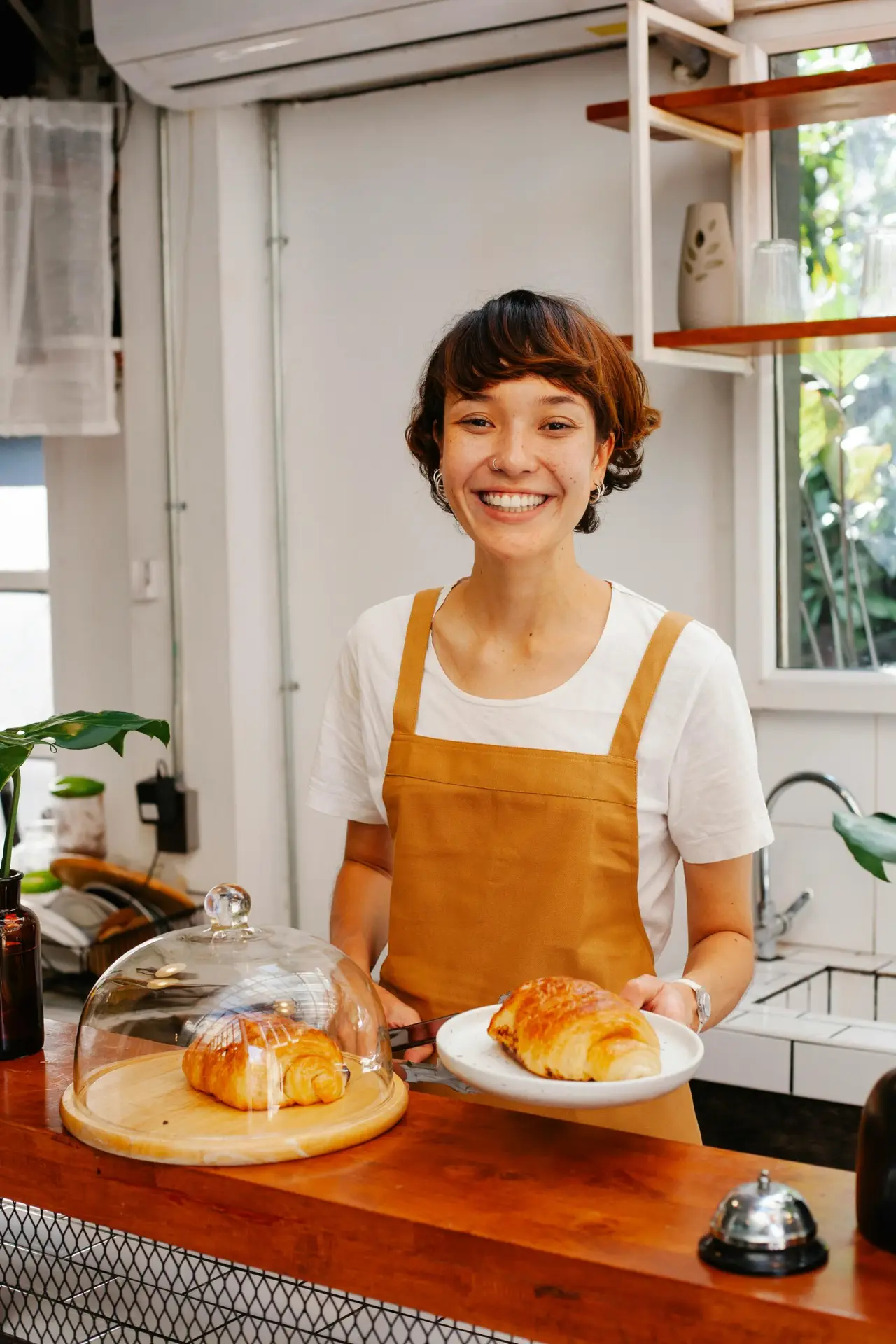 Charming waitress in apron serving croissant with a smile in a cozy café setting.